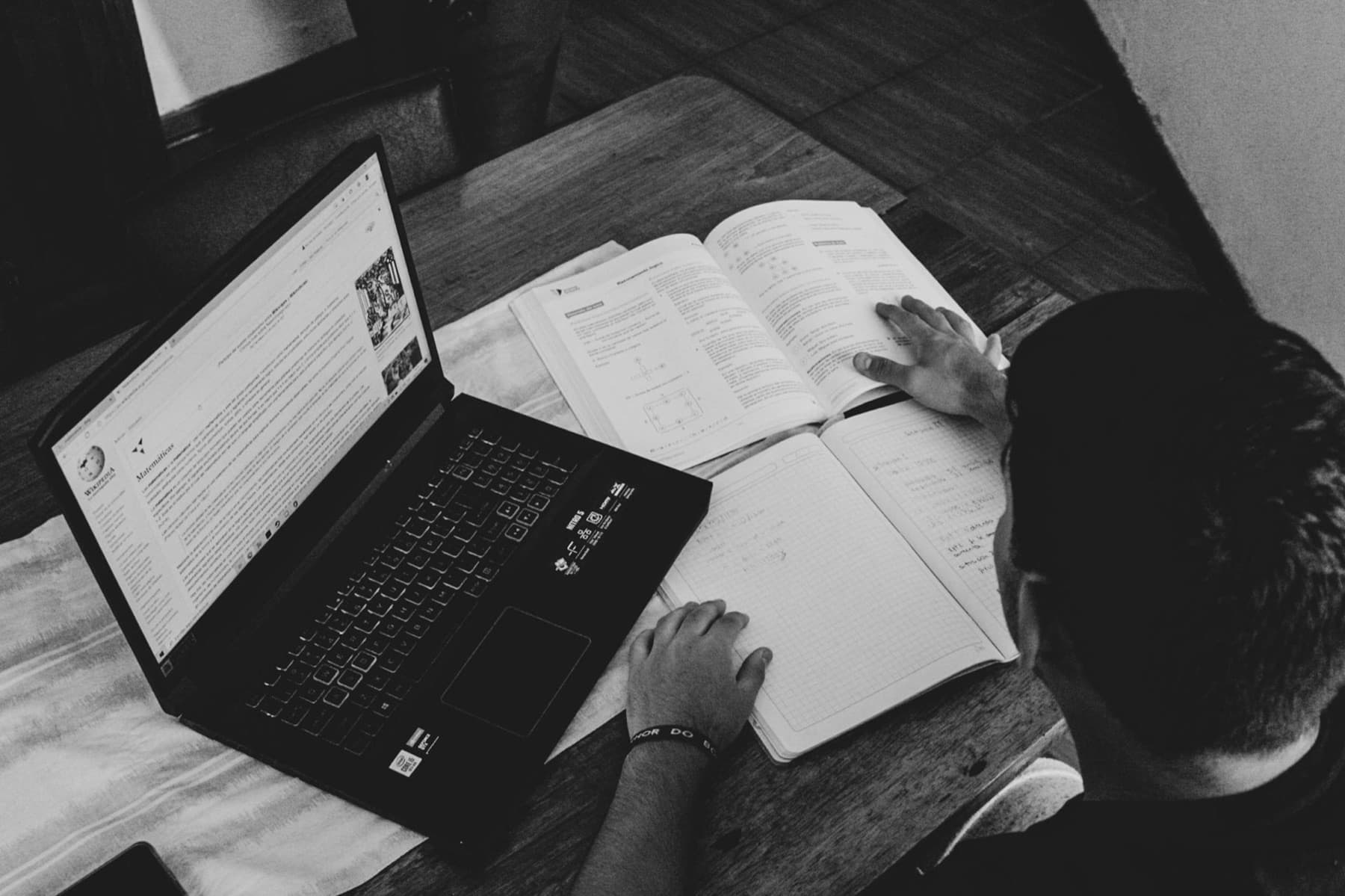 A black and white photo of a student reading a textbook beside a laptop